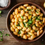 A bowl of cooked lupini beans garnished with parsley, with sea salt and lemon wedges on a rustic wooden table.