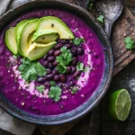 Close-up of a bowl of purple black bean soup with garnishes like avocado, cilantro, and lime on a rustic wooden table.